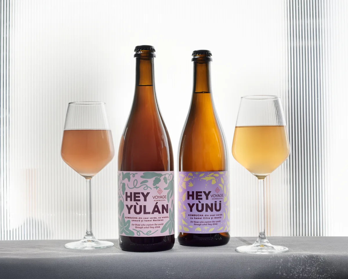 Două sticle de Voyage Kombucha — Hey Yùlán și Hey Yùnü — alături de pahare pline, pe un fundal lavandă